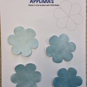 May include: A sheet of paper with the word "APPLIQUÉS" in blue, with four blue flower-shaped appliqués. A pencil sketch of a flower is in the upper right corner. The text "Ready to iron in place and stitch down" is below the word "APPLIQUÉS".