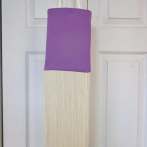 May include: A purple fabric tote bag with a long fringe of white yarn hanging from the bottom.