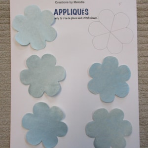 May include: A sheet of paper with six light blue flower-shaped appliqués. The paper also features a flower outline and the text "APPLIQUÉS" and "Creations by Melodie". The appliqués are ready to iron in place and stitch down.