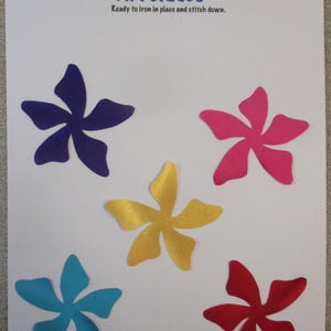 May include: A sheet of appliqués featuring five colorful flower shapes in blue, pink, yellow, red, and teal. The text "APPLIQUÉS" is displayed above the flowers, with the text "Ready to iron in place and stitch down."
