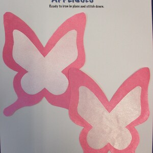 May include: Two pink butterfly appliqués with white centers are displayed on a white background. The text "APPLIQUÉS" is in blue, with the phrase "Ready to iron in place and stitch down." above the butterflies.