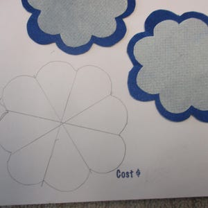 May include: A white paper with a pencil drawing of a flower with eight petals. The paper also has two blue felt flowers with white centers. The text "Cost $" is written on the paper.