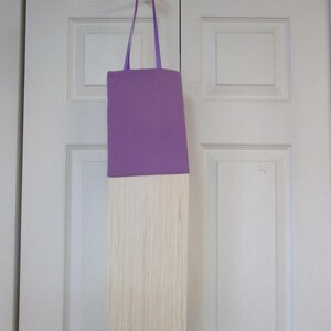 May include: A purple fabric tote bag with a long white fringe hanging from the bottom. The bag is hanging from a hook on a white door.