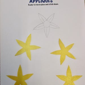 May include: A sheet of paper with the text "APPLIQUÉS" and "Ready to iron in place and stitch down." features a drawn outline of a flower and four yellow star-shaped appliqués. The paper is white.