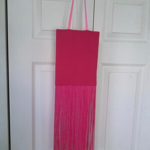 May include: A pink tote bag with a long fringe bottom. The bag has a pink fabric strap.