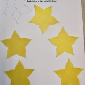 May include: A sheet of paper with the text "APPLIQUÉS" and a drawing of a flower. Five yellow star-shaped appliqués are arranged on the paper. The appliqués are ready to iron in place and stitch down.