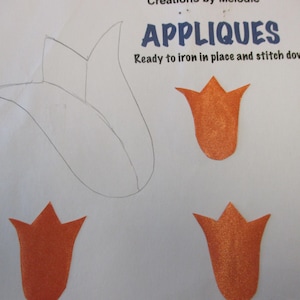 May include: A set of iron-on appliques featuring three orange tulip shapes. The appliques are ready to iron in place and stitch down. The text "APPLIQUES" is written in blue letters above the appliques. The text "Ready to iron in place and stitch down." is written in black letters below the appliques.