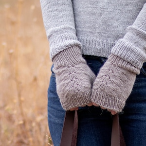Könnte beinhalten: Ein Paar braune gestrickte Fingerlose Handschuhe mit einem zarten Blatt-Muster. Die Handschuhe werden von einer Person getragen, die einen grauen Pullover und eine blaue Jeans trägt.