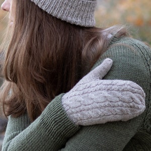 May include: A woman wearing a beige knitted hat and a green knitted sweater with a beige knitted mitten on her arm.