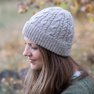May include: A gray knitted beanie with a cable knit pattern. The beanie is worn by a person with long brown hair.