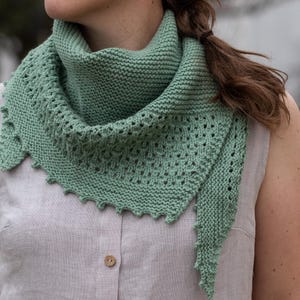 May include: A mint green knitted shawl with a triangular shape. The shawl features a textured knit pattern with a scalloped edge. The garment is worn around the neck and shoulders, offering a stylish and warm accessory.