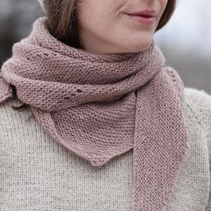 May include: A dusty rose-colored knitted scarf with a triangular shape. The scarf is wrapped around the neck and shoulders, with a portion of the scarf hanging down. The knitwear has a textured appearance and small holes near the top.