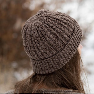 Könnte beinhalten: Eine braune Strickmütze mit geripptem Saum und strukturiertem Zopfmuster. Die Mütze ist aus einem weichen, warmen Material gefertigt, ideal für kaltes Wetter. Die Mütze wird von hinten gezeigt, wobei das Haar des Trägers sichtbar ist.