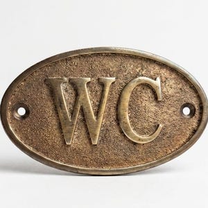 Könnte beinhalten: Ein ovales Messingschild mit den Buchstaben WC in erhabener, polierter Schrift auf einem texturierten, dunklen Hintergrund. Das Schild verfügt über zwei kleine kreisförmige Löcher an der linken und rechten Seite zur Befestigung.