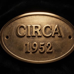 May include: An oval-shaped bronze-colored metal plaque featuring the raised text "CIRCA" on the top line and "1952" on the bottom line. The plaque has a textured surface and includes two circular mounting holes, one on each side of the text.