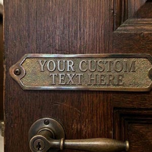 Puede incluir: Placa de puerta rectangular de latón con el texto "YOUR CUSTOM TEXT HERE" en fuente serif. La placa está montada en una puerta de madera oscura con un pomo de latón. La placa tiene una superficie texturizada.
