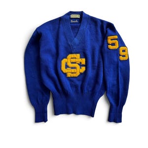 May include: A royal blue V-neck sweater featuring a large yellow felt letter patch on the chest and the numbers 5 and 9 in yellow felt on the left sleeve. The garment has a ribbed hem and cuffs, with a Roach brand label inside the neckline.
