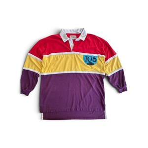 Pode incluir: Uma camisa de rugby de manga comprida com um design de blocos de cores, apresentando uma secção superior vermelha, uma faixa central amarela e uma secção inferior roxa. Possui gola branca, carcela de botões e um emblema azul com o número 106.