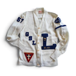 May include: A cream-colored knit cardigan featuring a V-neck, button-front closure, and ribbed cuffs. The garment displays dark blue felt lettering GSL and L, a number 57 patch, a triangular HI-Y patch, and a circular CP CHAMP 1958 patch.