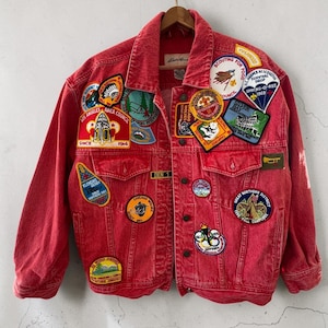 May include: A red denim jacket adorned with numerous patches. The patches feature various designs, including badges with text like "Los Angeles Area Council" and "Chicago Wolves." The jacket is hanging on a metal hook against a neutral background.