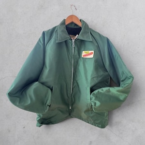 May include: Green zip-up jacket with a collared neckline and two front pockets. A rectangular patch with text is on the chest. The jacket is hanging on a wooden hanger.