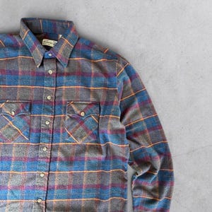 May include: A long-sleeve button-down shirt featuring a plaid pattern in shades of blue, grey, purple, and orange. The shirt includes a standard collar, a front button placket, and two chest pockets with buttoned flaps. A white label is visible inside.