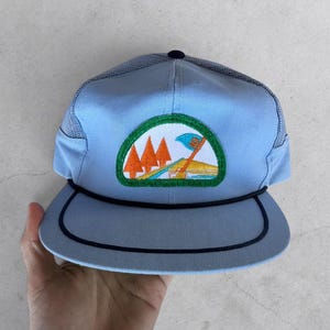 May include: A light blue baseball cap with a mesh back and a dark blue rope detail across the brim. The front features a semi-circular embroidered patch depicting orange pine trees, a blue flag, and a yellow landscape against a white background.