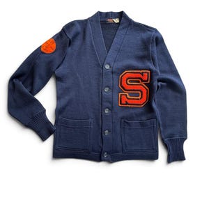 May include: A navy blue knit cardigan featuring a V-neckline, five front buttons, and two patch pockets. An orange letter S patch is on the left chest, and a circular orange patch with text is on the right sleeve. The cuffs are ribbed.