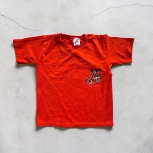 May include: Bright orange t-shirt with a small graphic of a tiger's face on the front. The shirt is short-sleeved and appears to be made of cotton. The graphic includes the text "Tiger Cubs 2003".