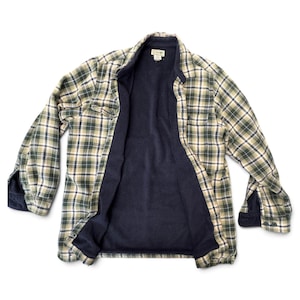 May include: A long-sleeve button-down shirt featuring a plaid pattern in shades of green, navy blue, and cream. The interior is lined with a solid navy blue fleece fabric. The shirt includes a collar, two chest pockets, and buttoned cuffs.