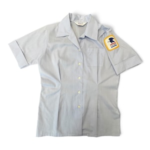 Puede incluir: Una camisa de color azul claro de manga corta con botones, cuello en punta y un bolsillo en el pecho. La manga izquierda tiene un parche rectangular con un gráfico de un águila azul y el texto U.S. MAIL en azul y rojo sobre fondo amarillo.