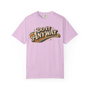 Puede incluir: Camiseta de color lila claro con la frase "DO IT ANYWAY" en una fuente retro. El texto está arqueado y tiene un diseño de arcoíris debajo. Pequeñas estrellas adornan el diseño.