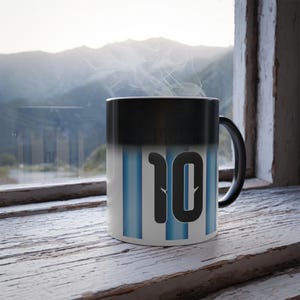 May include: A black and white color-changing mug with the number "10" in black. The mug has vertical blue stripes and is filled with a hot beverage, as indicated by the steam rising from the top. The mug is sitting on a wooden surface.