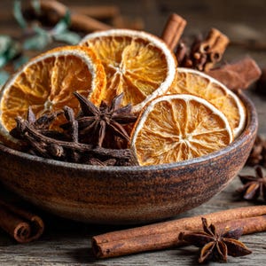 Puede incluir: Un cuenco texturizado de color marrón oscuro lleno de rodajas de naranja secas, anís estrellado entero y clavos de olor de color marrón oscuro. Varias ramas de canela están esparcidas alrededor de la base del cuenco, junto a anís estrellado.
