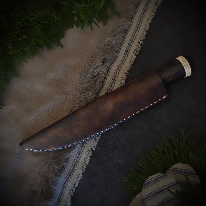 May include: A handcrafted knife with a dark wooden handle and a brown leather sheath. The sheath is stitched with white thread. The knife is resting on a dark surface with a patterned cloth and greenery in the background.