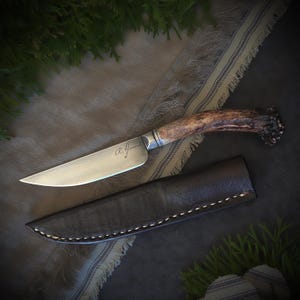 May include: A handcrafted knife with a polished steel blade and a handle made from antler. The blade is engraved with the name "R. Garrard". It is accompanied by a black leather sheath with white stitching.