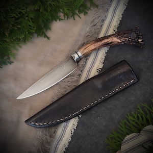 May include: A handcrafted knife with a polished steel blade and a handle made from natural antler. The knife features the engraved signature "R. Girard" and rests beside a dark brown leather sheath. The knife is approximately 9 inches long.