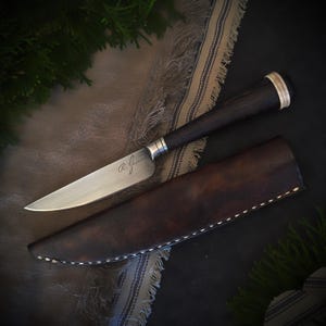 May include: A handcrafted knife with a dark wooden handle and a silver blade, resting in a brown leather sheath. The blade is engraved with the initials "R. Gasson". The knife is displayed on a patterned cloth.