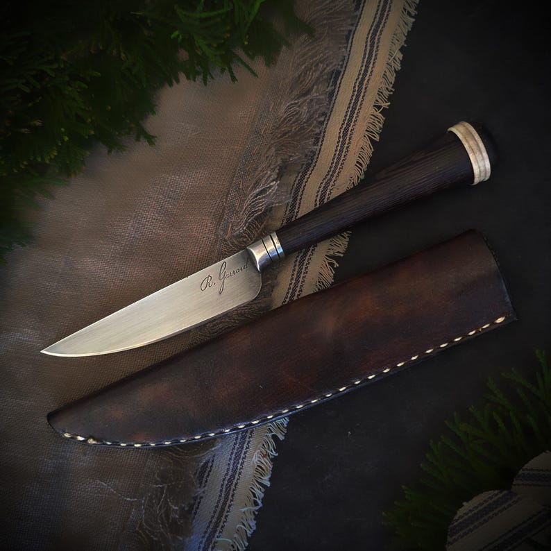 May include: A handcrafted knife with a dark wooden handle and a silver blade, resting in a brown leather sheath. The blade is engraved with the initials "R. Gasson". The knife is displayed on a patterned cloth.