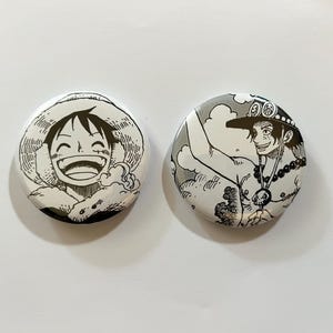 Könnte beinhalten: Zwei runde, weiße Buttons mit schwarz-weißen Manga-Illustrationen. Ein Button zeigt einen lächelnden Charakter mit Strohhut, der andere einen Charakter mit Hut und Halskette, beide aus der Anime-Serie.