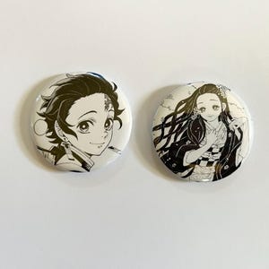 Könnte beinhalten: Zwei runde, weiße Buttons mit schwarz-weißen Anime-Charakter-Illustrationen. Ein Button zeigt einen männlichen Charakter mit dunklem Haar, der andere einen weiblichen Charakter mit langem, fließendem Haar.