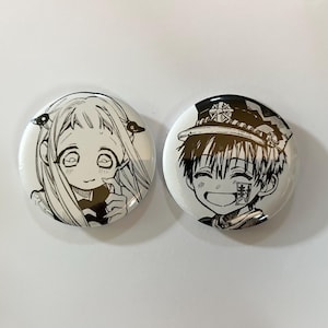 以下が含まれることがあります： 白地に黒と白のアニメキャラクターのイラストが描かれた丸いバッジが2つ。1つは長い髪の女の子、もう1つは帽子をかぶった男の子が描かれています。これらのバッジは、アニメシリーズのファン向けである可能性があります。