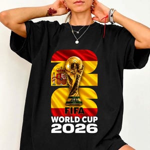 Może przedstawiać: Czarny t-shirt z grafiką trofeum Mistrzostw Świata FIFA umieszczoną na tle hiszpańskiej flagi. Projekt zawiera napis FIFA WORLD CUP 2026 pogrubioną białą czcionką. Trofeum jest złote z zieloną podstawą, a flaga przedstawia czerwone i żółte poziome pasy z hiszpańskim herbem.