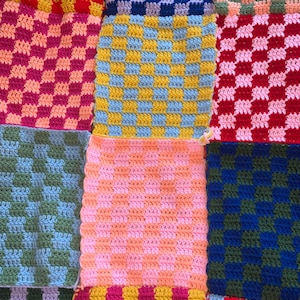 Może przedstawiać: Patchwork z szydełkowych kwadratów w różnych kolorach, w tym czerwonym, różowym, niebieskim, zielonym i żółtym. Każdy kwadrat ma teksturowany, w kratkę wzór. Ręcznie robiony materiał prezentuje żywy, wielokolorowy design.