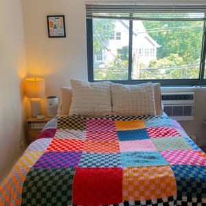 May include: A handmade crochet blanket featuring a vibrant patchwork design of multi-colored checkered squares in shades of red, blue, yellow, green, pink, and orange, finished with a black and white checkered border along the bottom edge.