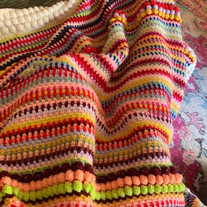 May include: A multi-colored crochet blanket featuring horizontal stripes in various vibrant hues including red, orange, yellow, green, blue, and purple. The textile incorporates textured bobble stitch rows and a thick, cream-colored border edge.