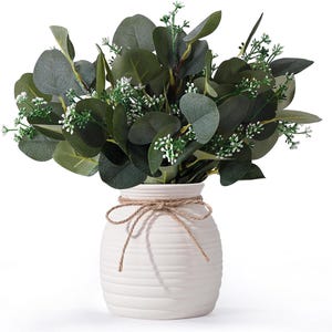 May include: A white ceramic vase with horizontal ridges and a tied jute twine bow around the neck, filled with a dense arrangement of dark green eucalyptus leaves and small white flower sprigs, creating a full, rounded botanical display.