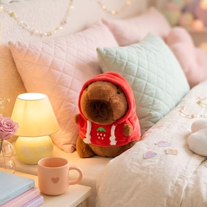 May include: A plush brown capybara wearing a red hoodie with a strawberry design. A small, yellow lamp and a pink mug with a heart are on a nightstand. Soft pink and green pillows are in the background.