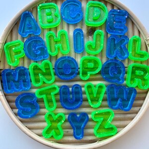 May include: A set of alphabet letters in blue and green hues, arranged on a circular, woven tray. The letters are made of a translucent material with glitter. The letters are a fun and colourful educational tool.