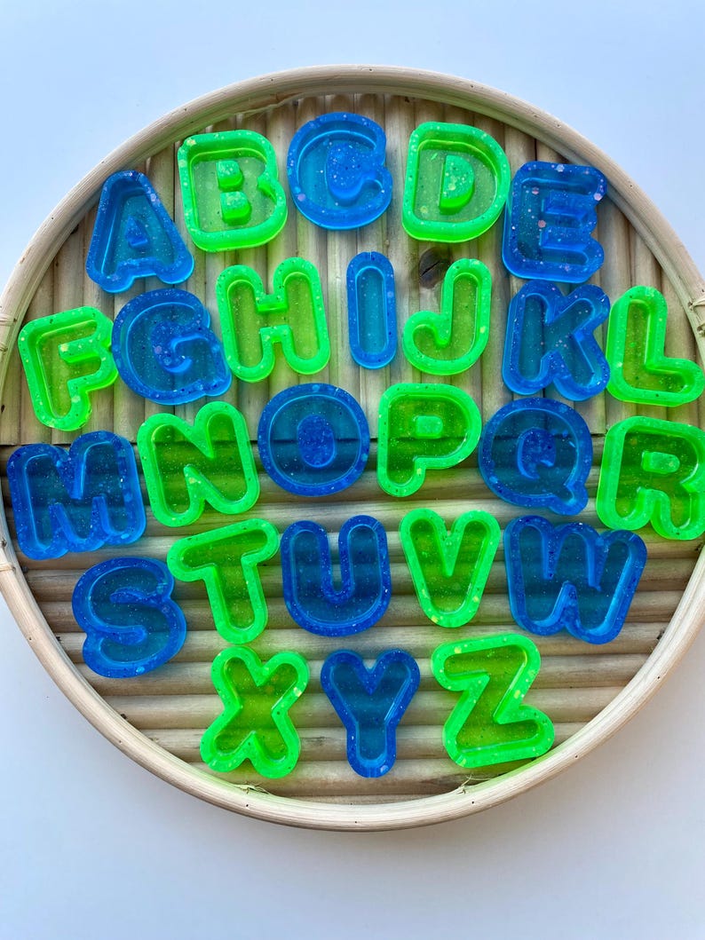 May include: A set of alphabet letters in blue and green hues, arranged on a circular, woven tray. The letters are made of a translucent material with glitter. The letters are a fun and colourful educational tool.